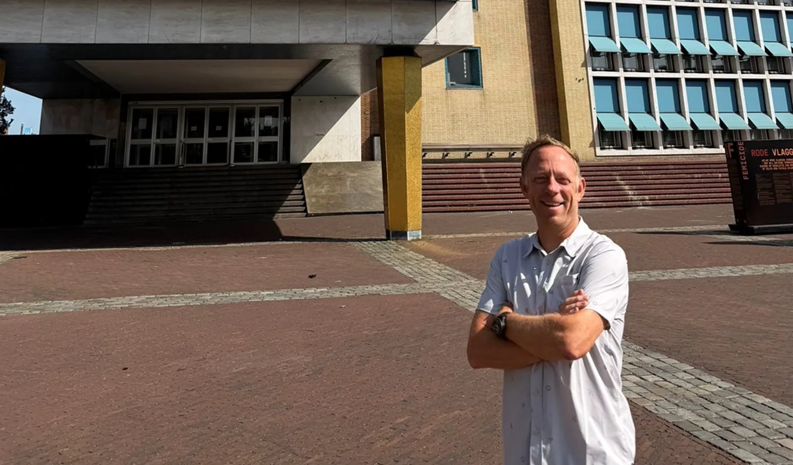 Man met gekruiste armen staat glimlachend op een plein voor een gebouw met blauwe raamkozijnen, sfeerbeeld bij het artikel 'je hebt mensen nodig die echt inhoudelijk meedenken